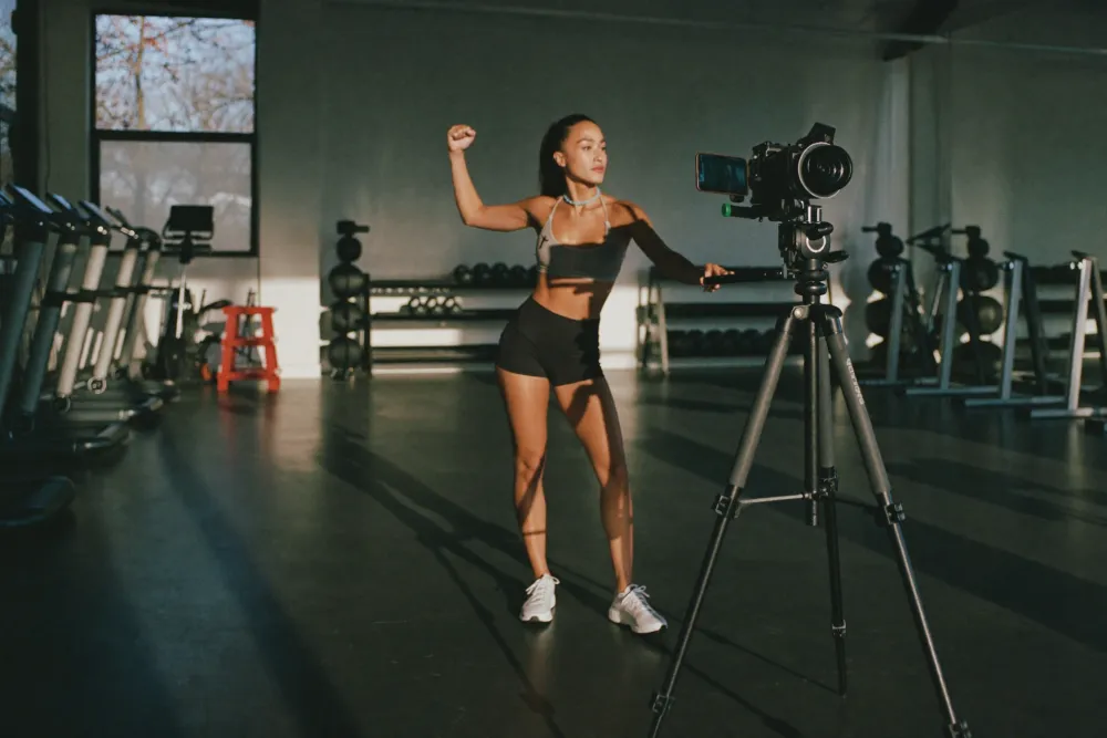 A woman films herself on a tripod-mounted camera in an empty gym, mid-move between the weight racks