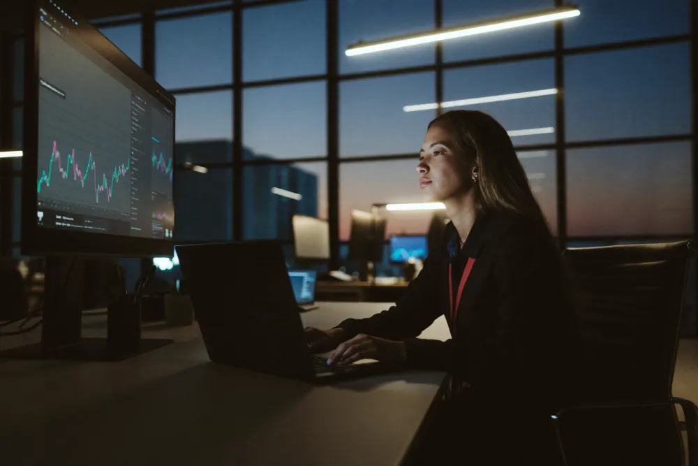 A woman at a laptop in a dark office at night, a wall of trading charts glowing beside her