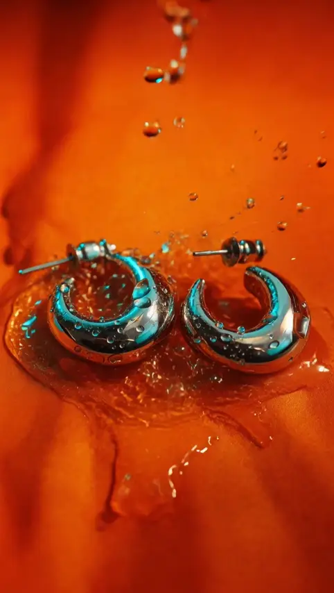 Chunky silver hoop earrings sit in beads of water on rippled orange fabric