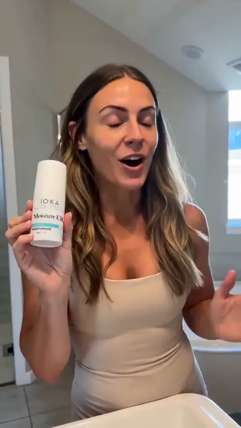 A woman holds a moisturiser bottle to camera, dispenses it and presses it into her cheeks