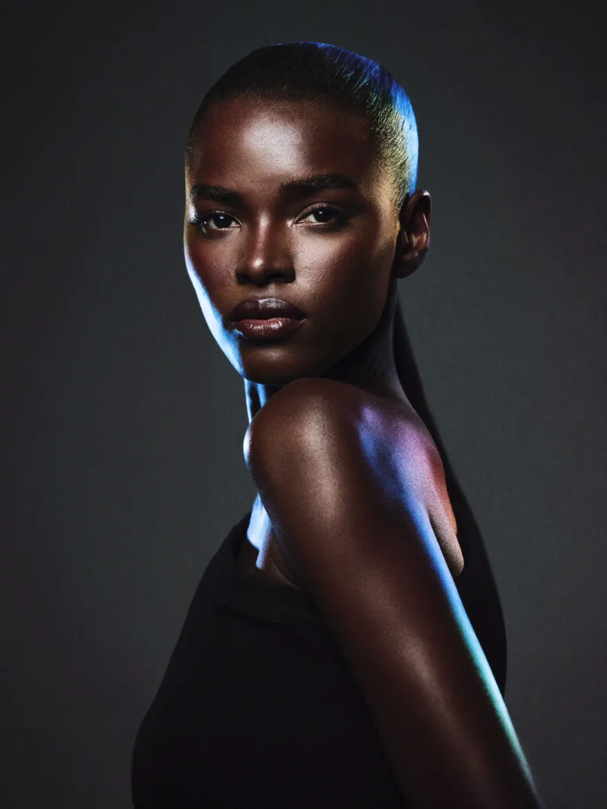 A woman with a shaved head in a black top, lit from the side with a violet rim light against black